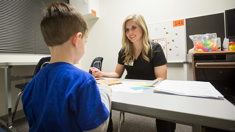 A speech therapy student sits at a table across from a young child during a therapy session.