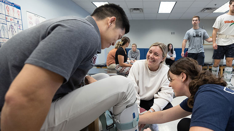 Two physical therapy students helping another student put on prosthetic footwear.