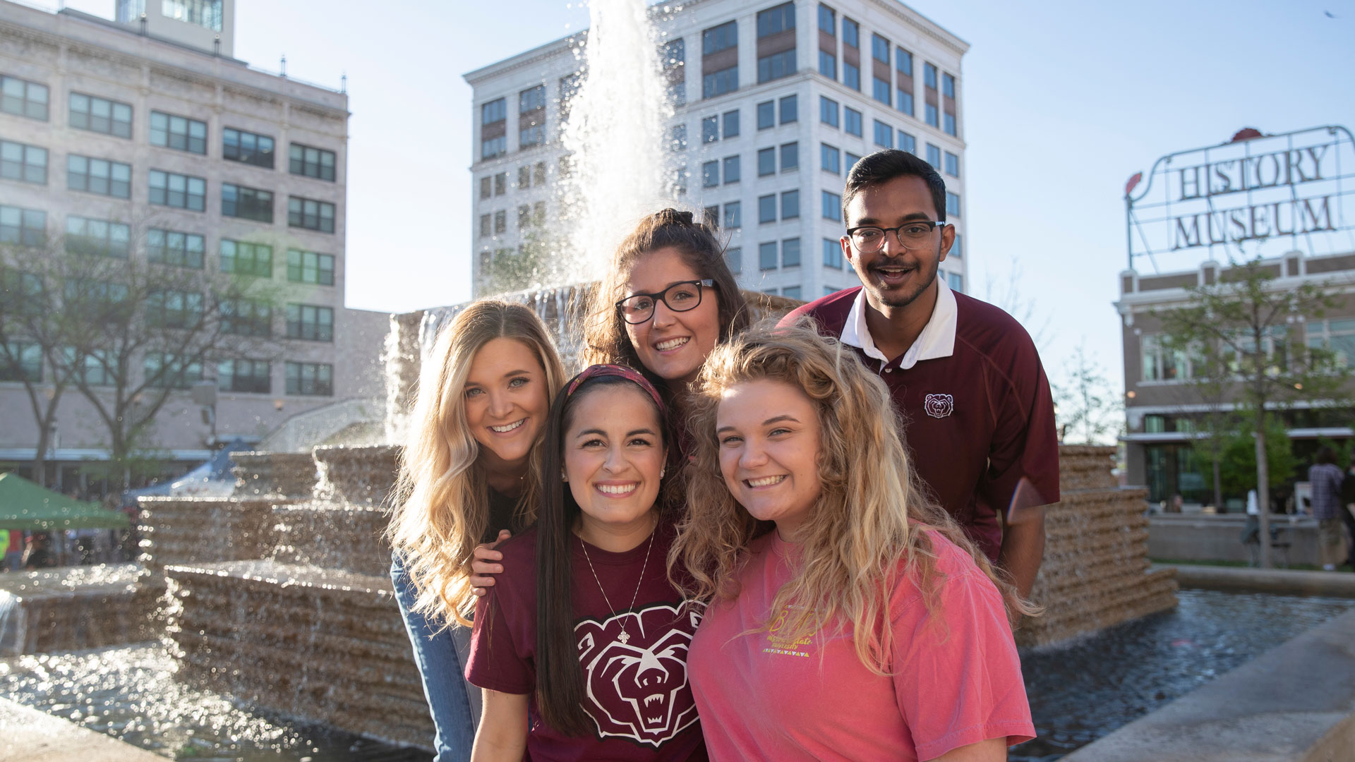 Five smiling students in front of the downtown fountains on Park Central Square.