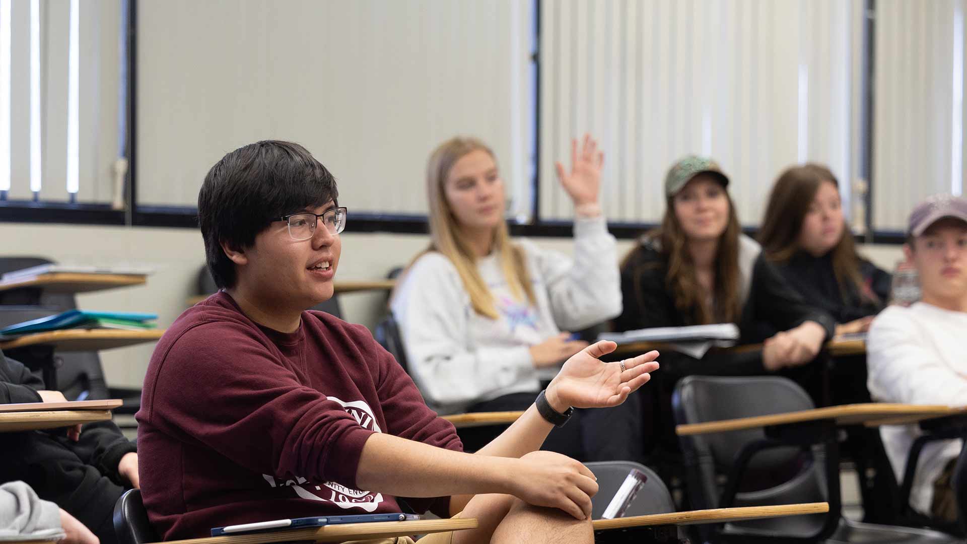 A communication student participates in a class discussion while another student raises her hand in the background.