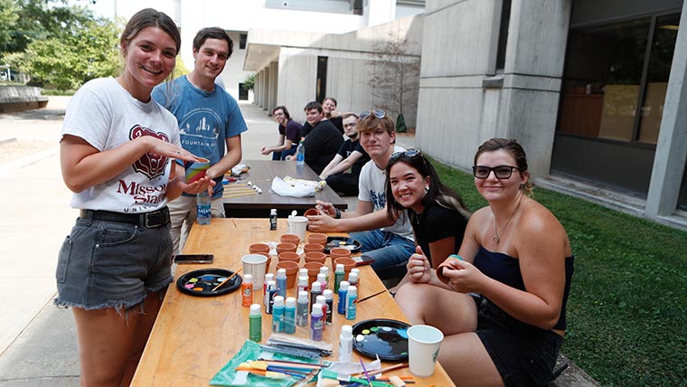 Students from the STEM living-learning community smiling at the camera during a social event. Arts and craft supplies, including paint, are spread across a big table.