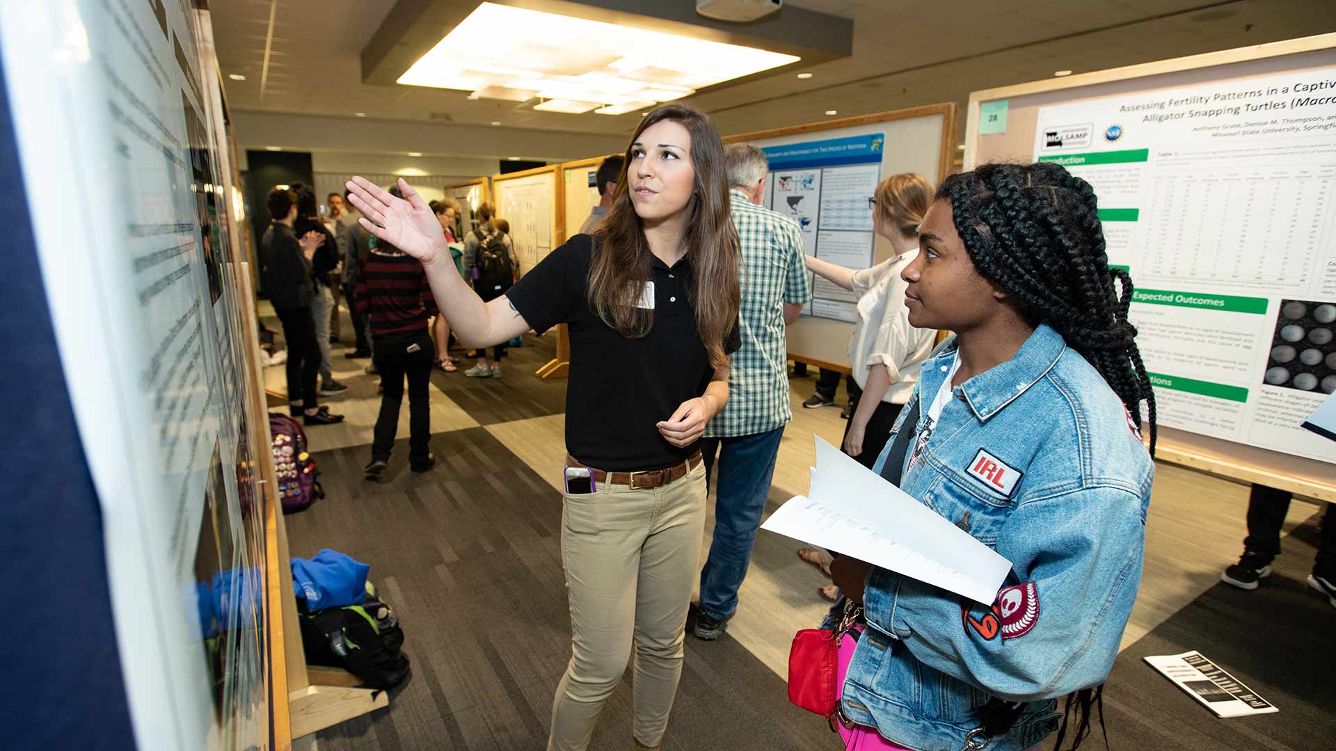A student explains her research presentation to another student during the 2019 College of Natural and Applied Sciences Undergraduate Research Symposium.