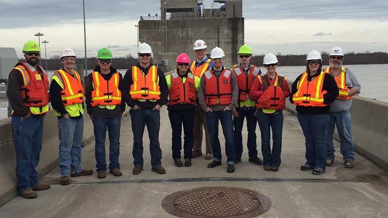 Missouri State engineering students posing for a photo at the Olmsted Lock and Dam facility in Illinois.  The students and their tour guides are all wearing orange safety vests and hard hats.