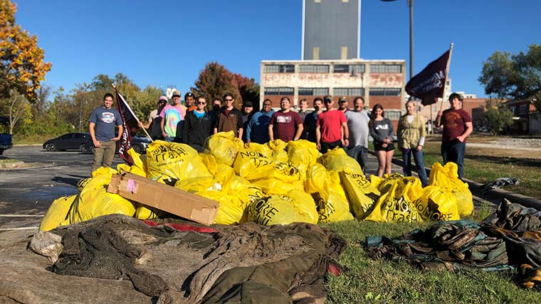 Yellow trash bags filled with litter after a community cleanup event. In the background is a big group of engineering students, two of which are holding Missouri State University flags.