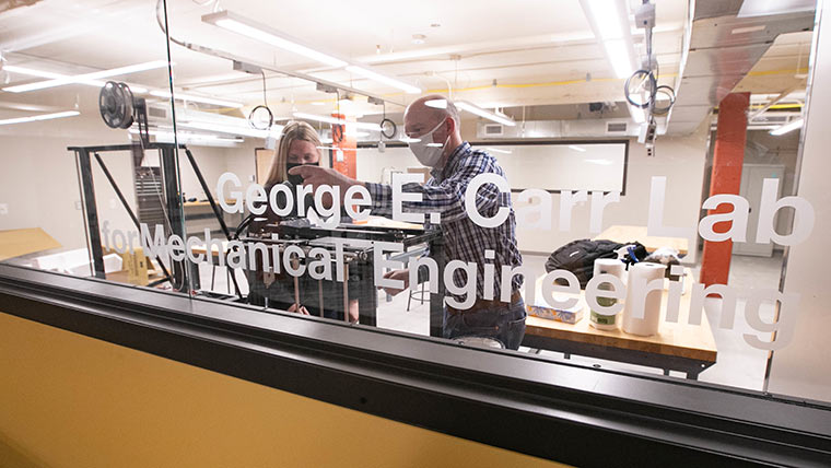 An other-side-of-the-glass view of two people working in an engineering lab. The lab's name, George E. Carr Lab for Mechanical Engineering, is displayed on the glass.