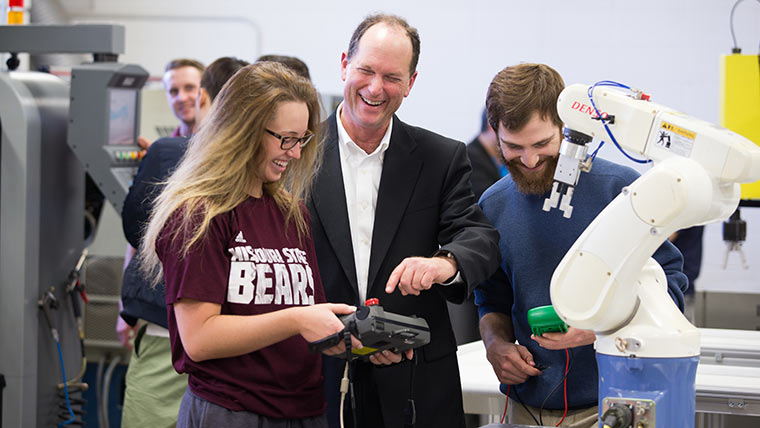 Two students and a professor using a controller to operate a robotic arm. All three are laughing and having fun.