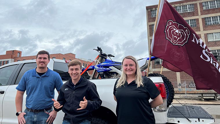 Three engineering students posing for a photo in front of a pickup track. In the truck bed  are a go-cart and a maroon Missouri State Bears flag