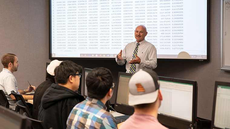 Dr. Randall S. Sexton, professor of information technology and cybersecurity, speaking to students in class. In the background is a data chart shown on a projector screen.