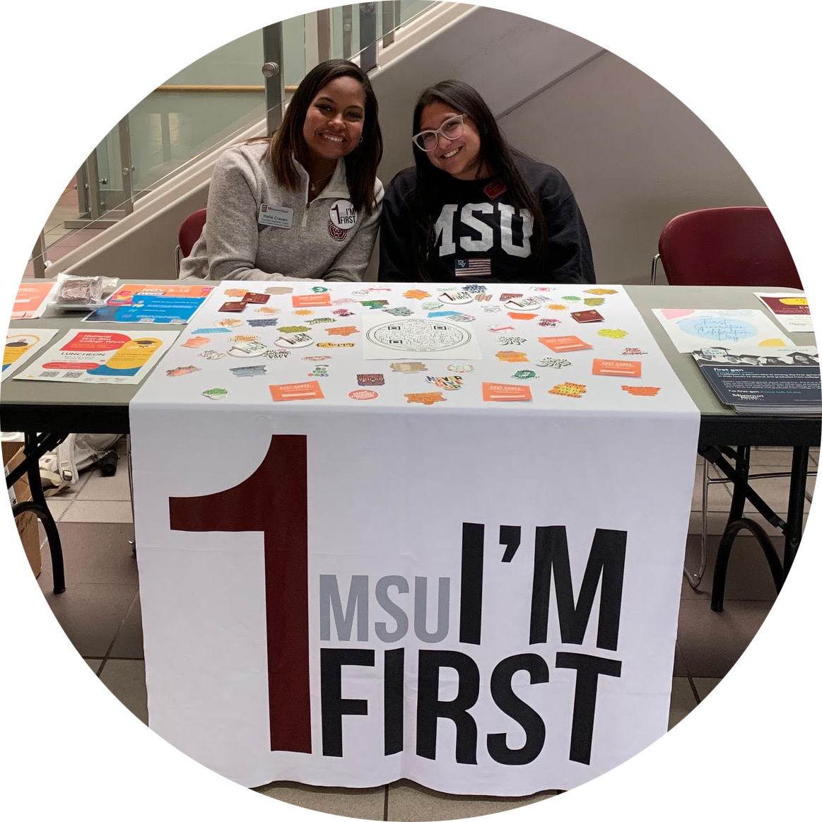 Two students smile into the camera. They are sitting behind a table with various materials on it and a banner that says 1 MSU I'm First