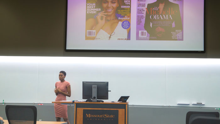 Maya Sudduth speaking while projection screen displays magazine covers that relate to her senior project.