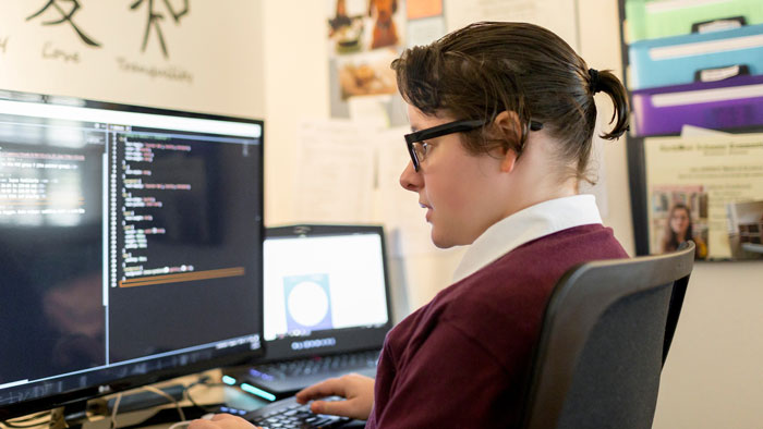 Madison Estabrook sitting at computer desk with coding information displayed on the monitor.