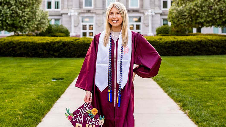 A Bachelor of Social Work (BSW) graduate standing in front of Carrington Hall while wearing a maroon commencement gown. She's holding a maroon cap that's decorated with flowers and the phrase 'Be the change: BSW'
