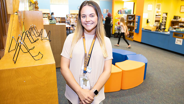 A social work student posing in the library of an elementary school. Students are moving around in the background.