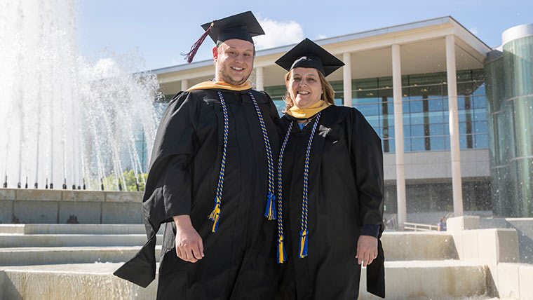 A pair of Master of Social Work (MSW) graduates wearing their commencement cap and gown while standing in front of the Hammons Fountain.