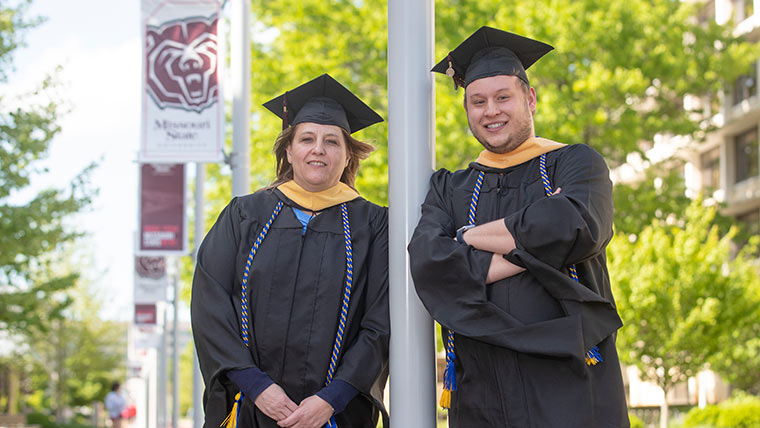 Master of Social Work (MSW) graduates Jayma and Ethan Potter posing on a sunny day on campus. Both are wearing a commencement cap and gown.