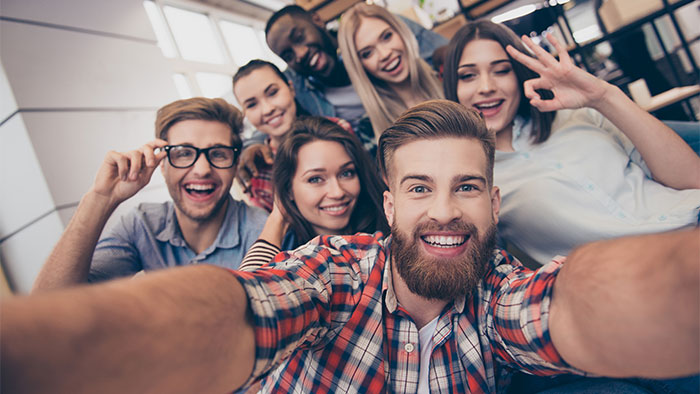 A group of seven smiling students crowded together to take a selfie; it looks like they are in a library