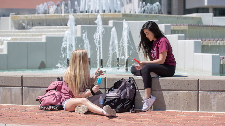 Two female students eat popsicles as they sit by the fountain