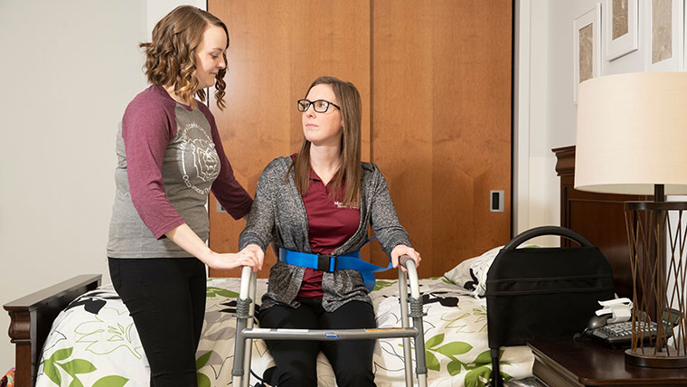OT student  helping another student using a walker get up from bed.