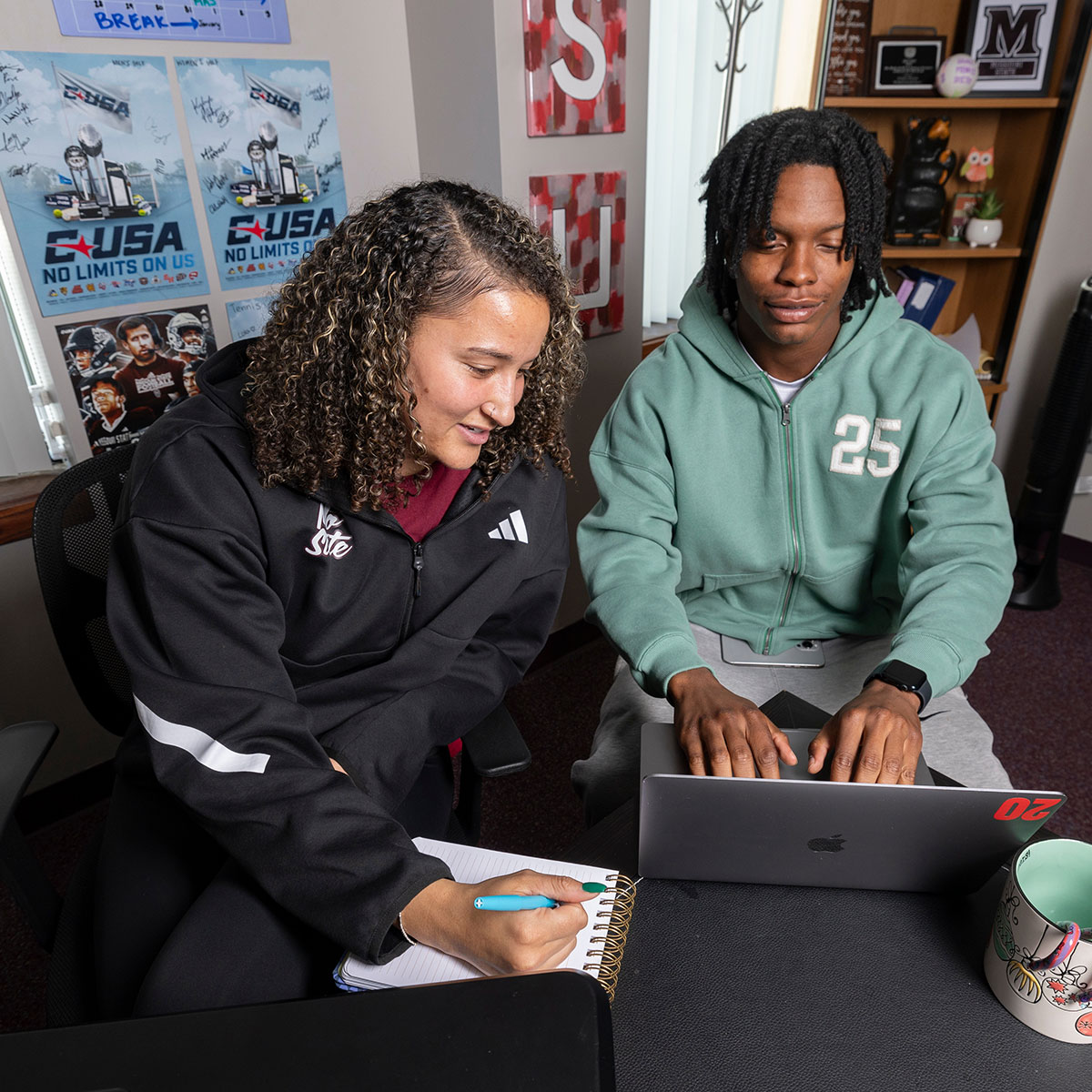 An advisor brings out a notepad and pencil to draw up an academic plan for her advisee, as he types on his laptop.