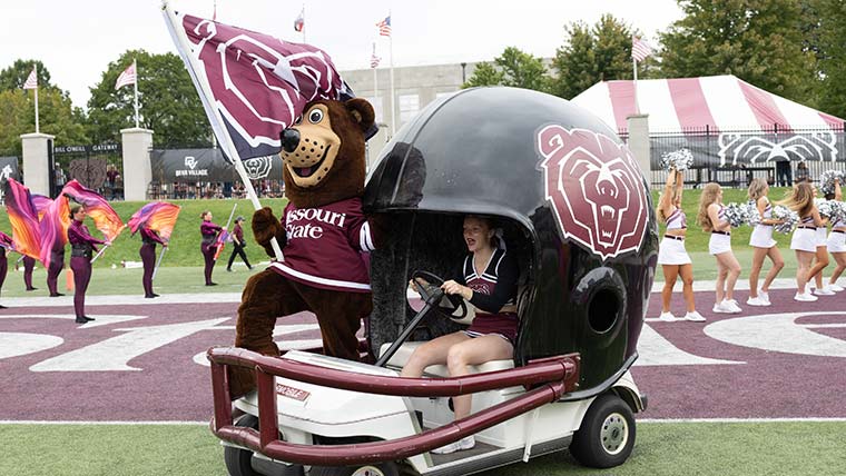 Boomer Bear holding a flag and standing in the football cart as a cheerleader drives the cart onto the field before a home football game.