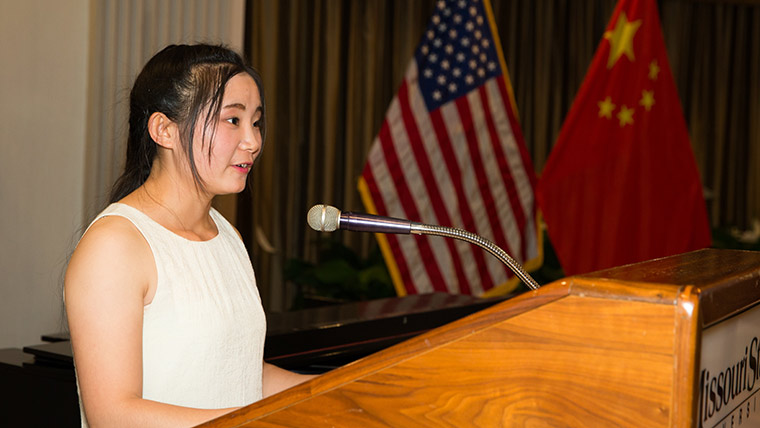 A Missouri State student from China speaks to an audience. The United States and China flags are seen in the background.