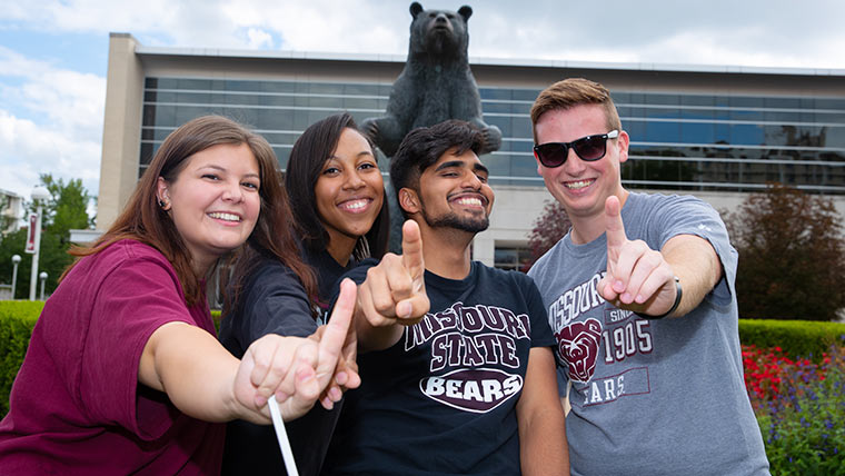 Four students in Bearwear pose together in front of the Bear statute outside Plaster Student Union.