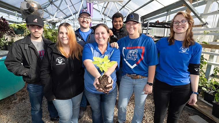 West Plains ag students pose for a group photo in a greenhouse. Their teacher is in the center and holding a plant.