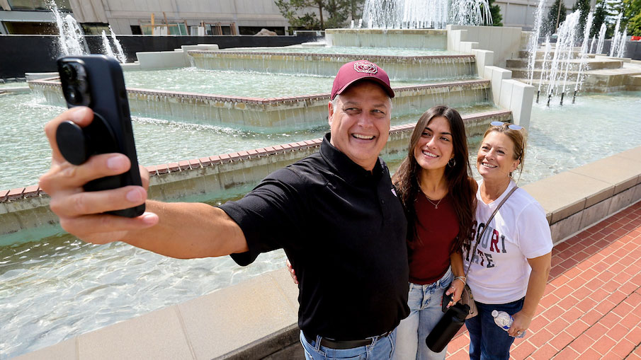 A dad taking a selfie of his daughter and wife in front of the fountain