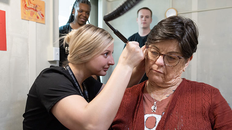 An audiology student uses a medical device on a patient's ear. Her professor and classmate observe in the background.