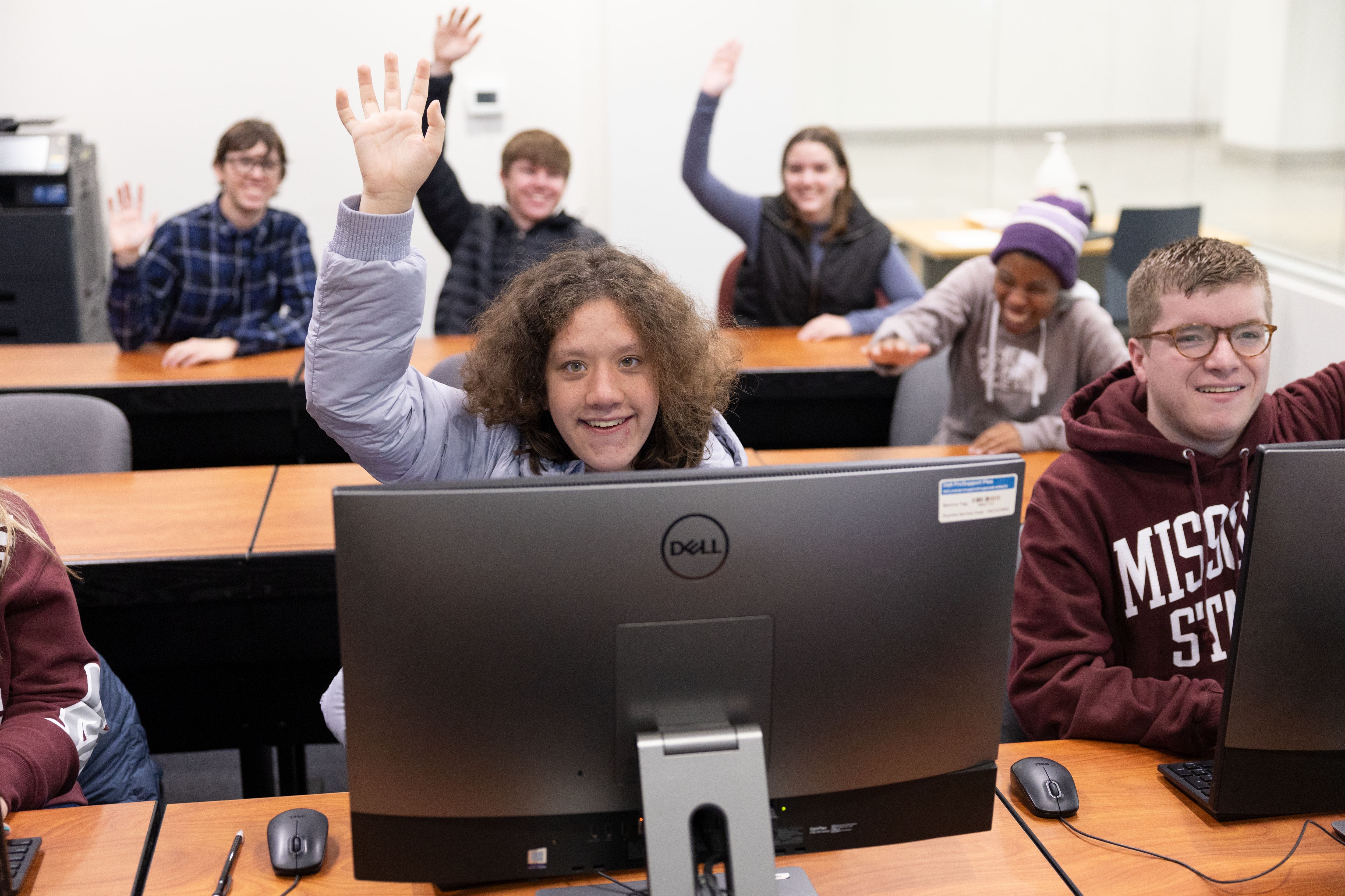 Students sitting in the computer lab with hands raised and smiling