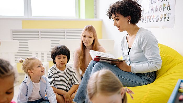 A woman and kids in a classroom, the woman is reading a children's book and smiling