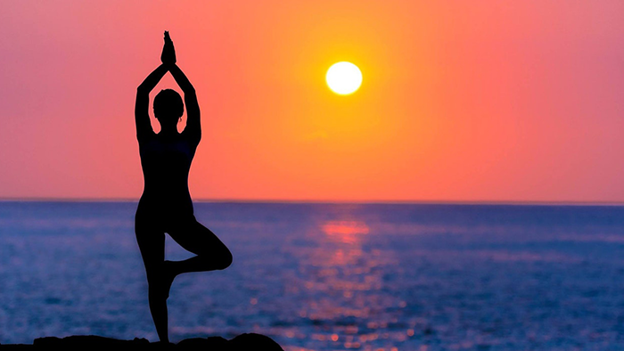 A woman does yoga, silhouetted against an ocean sunset.