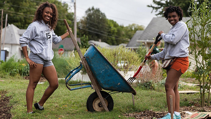 Two young women move mulch from a whellbarrow onto a garden while smiling at the camera