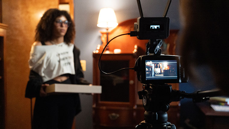 An actress carries a box of doughnuts onto the set as she is being filmed by a camera.