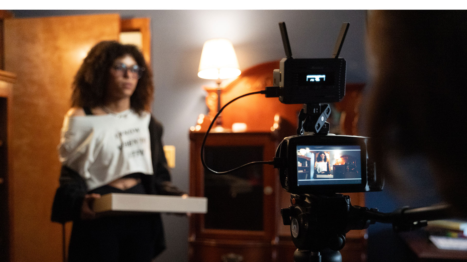 An actress carries a box of doughnuts onto the set as she is being filmed by a camera.