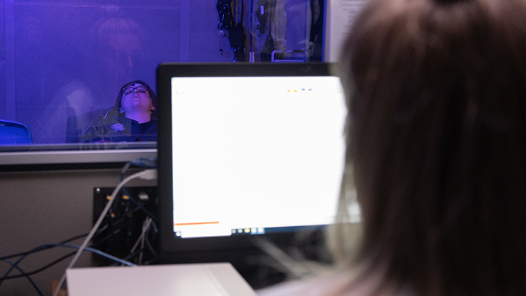 An over-the-sholder view of an audiology student reading data on a computer screen. Another student is laying in a chair across from her, behind a glass screen, as the testing is conducted on her.