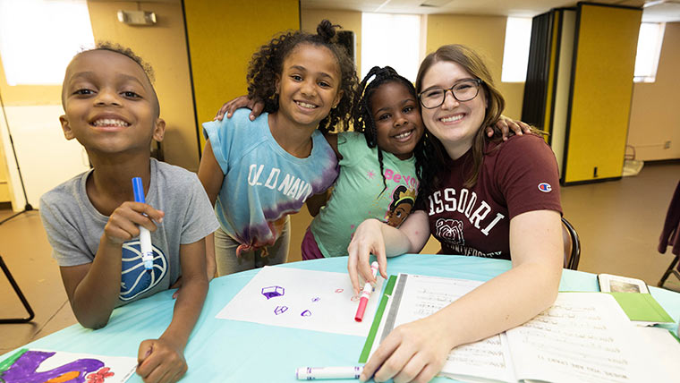 A Missouri State student and three young children smile for a group photo during Ujima Literacy Night at a local elementary school.