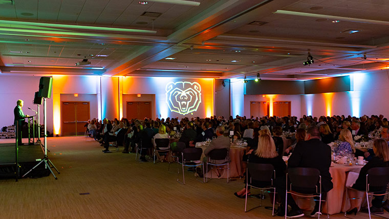People seated in a banquet hall during the Bears of Distinction Awards. A large Bearhead logo is on display in the background.