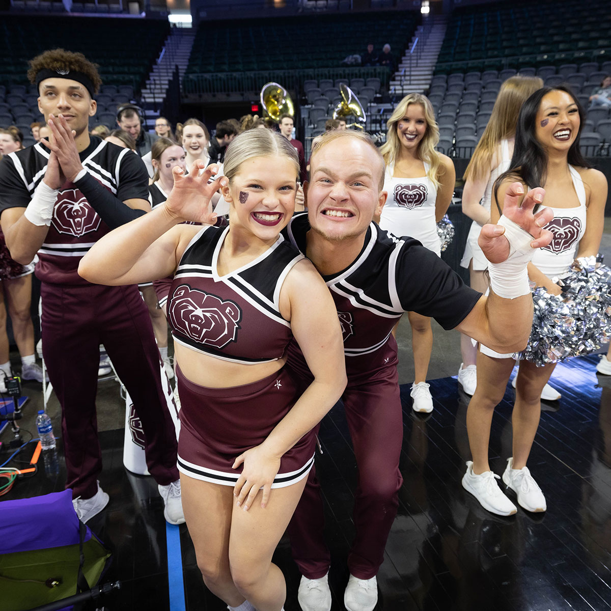 Two cheerleaders pose for a picture, giving a growling face while making a bear claw gesture with their hands. Other MSU cheerleaders and Sugar Bears smile in the background.