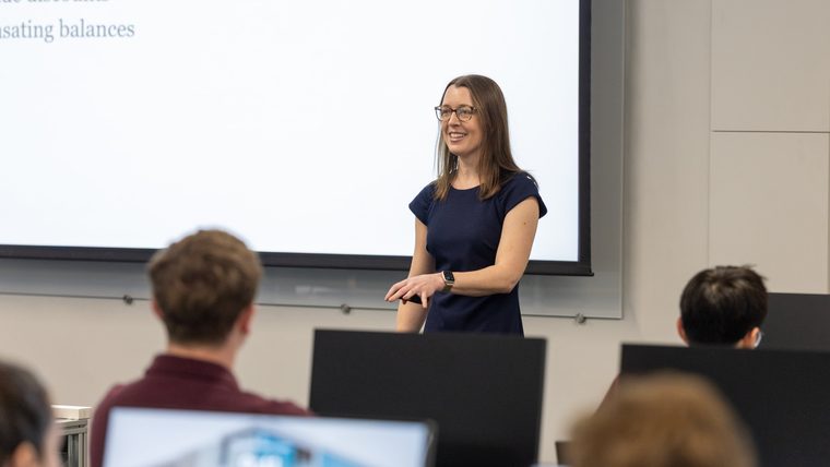 A finance instructor lectures to students in front of a projector screen.