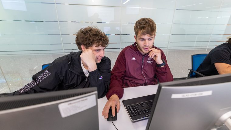 Two Missouri State students in Bearwear work together at a computer in the lab.