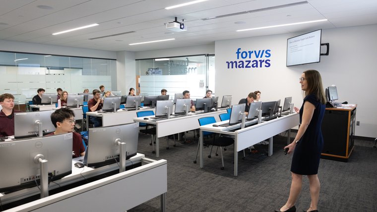 An instructor lectures to students at computer workstations in the Forvis Mazars Financial Trading Lab.