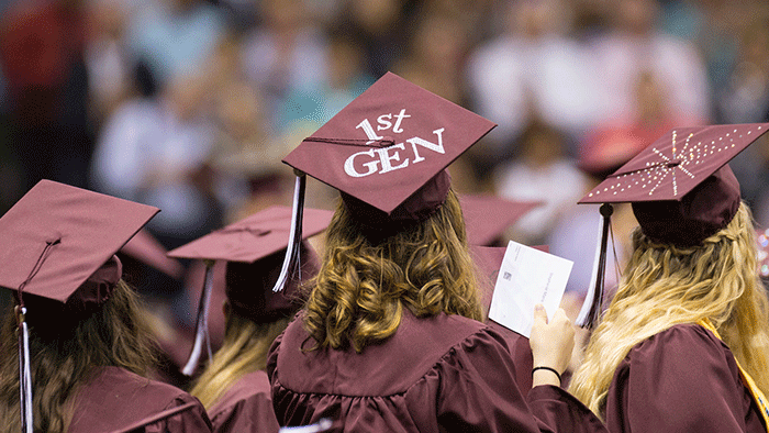 A view from behind of three MSU students in graduation caps and gowns. The cap for the student in the middle says 1st Gen.