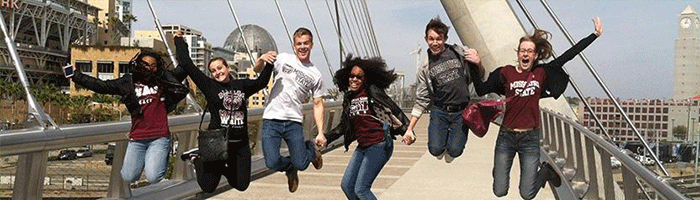 Six students in MSU clothing are in midjump on a pedestrian bridge with San Diego in the background