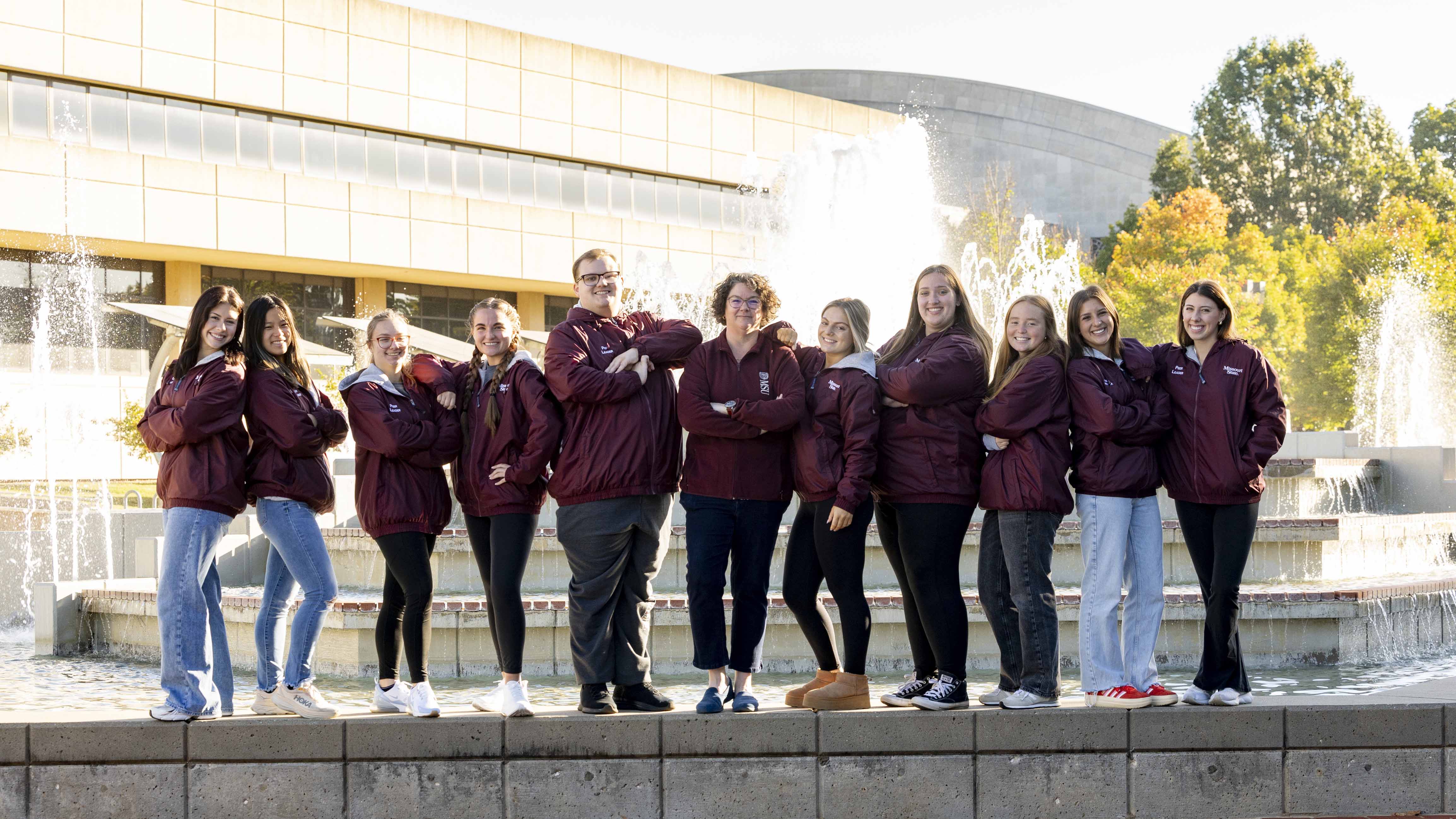 A group of smiling people in matching maroon jackets stand in a line in front of a fountain