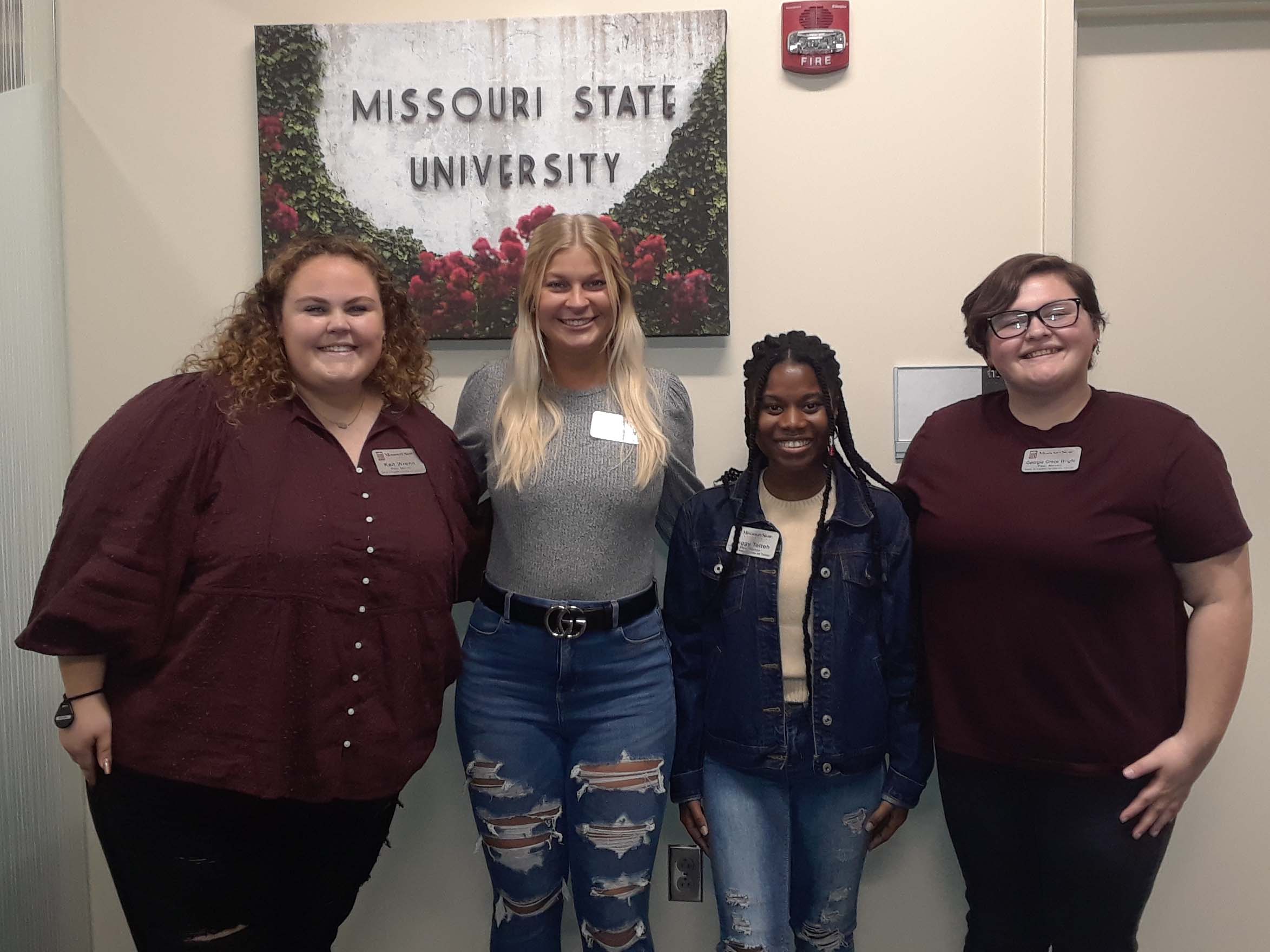 Four smiling peer mentors. On the wall behind them is a canvas print with the words Missouri State University.