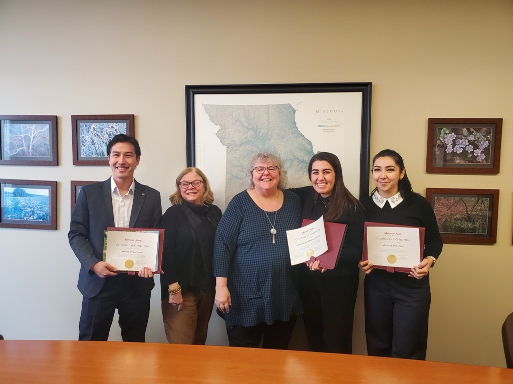 Five people smiling together in an office while holding certificates of completion in front of a framed map of Missouri.