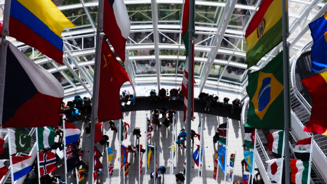 An overhead view looking down into a bright glass atrium with international flags hanging from tall poles and people gathered in the open space below.
