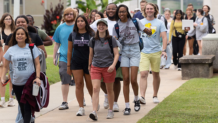 A large group of smiling students walking together outside on a campus sidewalk.