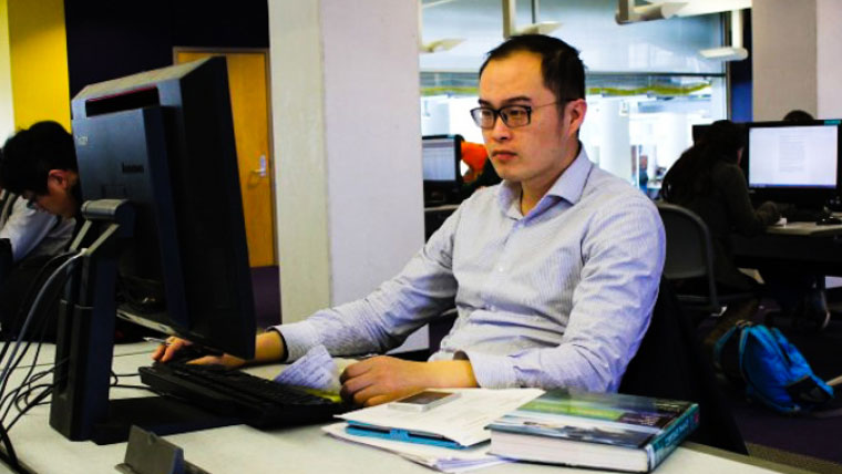 A young man wearing glasses working at a computer station in a campus library or computer lab